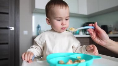 Sakin, tapılası bir bebek yemek masasında oturuyor. Çocuk annesinin tam kaşık vermesini bekliyor. Kapatın. Bulanık arkaplan.