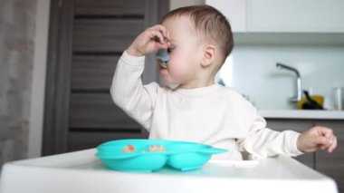 Ağzında kaşık tutan sevimli bir çocuk. Bebek, yemek masasında mavi tabaktan yiyor. Düşük açı görünümü.