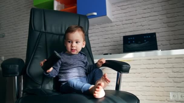 裸足のアクティブなスマイリーベイビー男の子は オフィスの椅子に座っています テレビのリモートと椅子で彼の手を叩く遊び心のある子供 — ストック動画