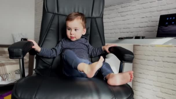 大きな革の椅子に快適に座っている小さなベビーボス かわいい少年はテーブルからテレビをリモート取る — ストック動画