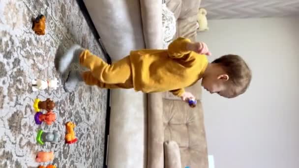 黄色いスーツを着たかわいいコーカサスの赤ん坊は 床に玩具の動物を置きました 子供は遊ぶおもちゃを選択する 垂直ビュー — ストック動画