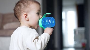 Bebek emzik ile mavi bir şişeden su içiyor. Yan görüş. Bebek merakla yukarı bakıyor. Bulanık arkaplan.