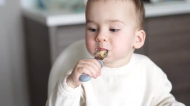 Beyaz, tatlı çocuk yavaşça kaşıktan yulaf lapası yiyor. Sevimli bebek dikkatlice yemeğini çiğniyor. Kapat..