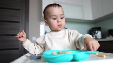 Beyaz kazaklı sevimli beyaz bebek evde yemek yiyor. Çocuk mavi tabakla masada oturuyor ve annesi oğluna tam kaşık veriyor. Bulanık arkaplan.