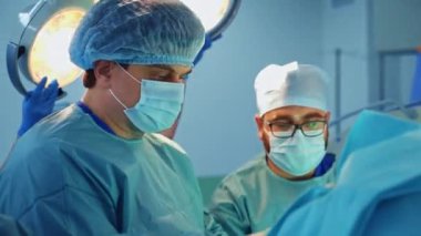 Ameliyatta işbirliği yapan iki yetişkin erkek cerrah. Hemşire arka planda ışığı tamir ediyor..