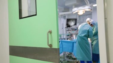 Modern cerrahi odasında ameliyat yapan iki erkek doktor. Açılmış kapıdaki odaya bak..