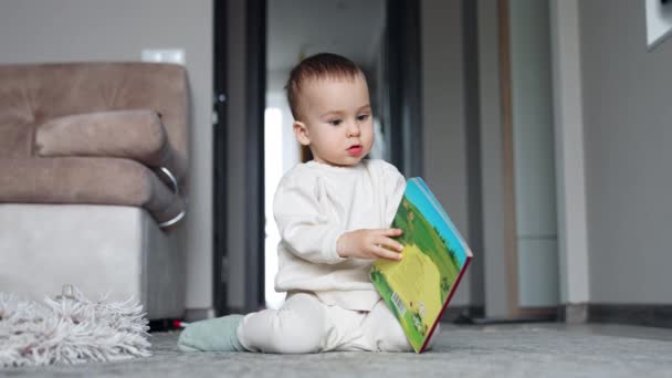 いい子が本を持って床に座っている 愛らしいトッドラーは本を開こうとするが失敗する — ストック動画