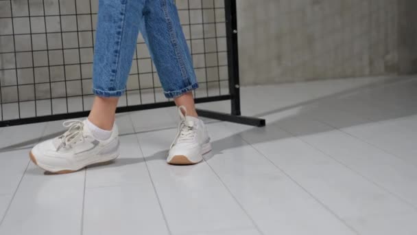 女孩转身展示舒适的白色运动鞋 穿蓝色牛仔裤的女人穿着时髦的鞋子走路 背景为黑色格栅 — 图库视频影像
