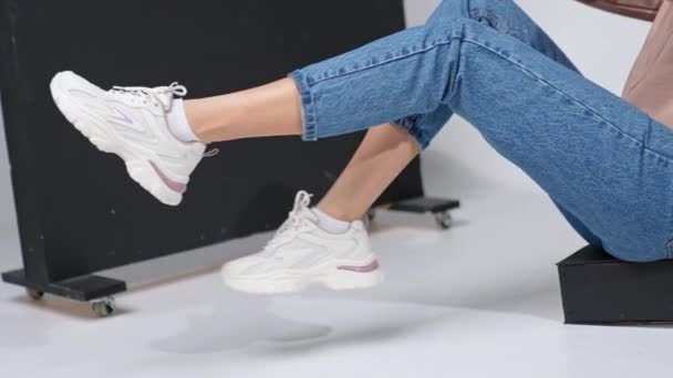 穿着牛仔裤和白色时髦运动鞋的女孩坐在台阶上挥动着脚 工作室里的现代鞋类示范 — 图库视频影像