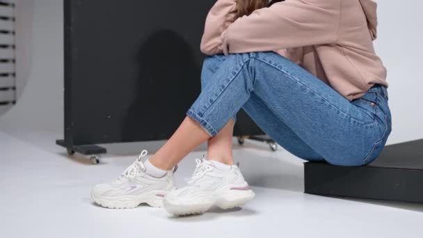 剪下了一个女孩坐在黑色台阶上的图像 工作室里的牛仔裤 运动衫和白色运动鞋模型 — 图库视频影像