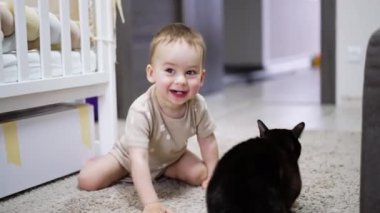 Sevimli gülümseyen çocuk siyah bir kediye doğru sürünüyor. Bebek yanağını kediye koyar ama evcil hayvan ayağa kalkar..