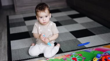 Beyaz bebek kameraya odaklanmış. Çocuk elinde oyuncağı tutarak yerde oturuyor..