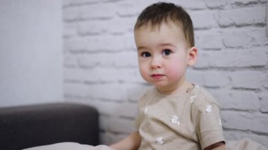 Güzel beyaz çocuk yatağın üzerine oturur. Bebek heyecanla yastığını sallıyor. Neşeli ruh hali.