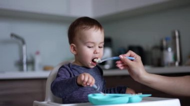 Anne oğlunu kaşıkla besliyor. Çocuk çatalıyla tabağa vururken anne çocuğun ağzını temizlemek için peçete kullanıyor..