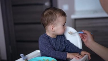 Beslenme masasında oturup dönüşen sevimli bir bebek. Annem kaşığı bir çocuğun ağzına sokuyor. Bulanık arkaplan.