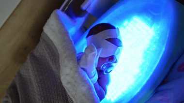 Gözlerinde bandaj ve emzik olan yeni doğmuş bebek ultraviyole lambada yatıyor. Çocuk ellerini göğsüne koydu. Sarılığa karşı terapi.