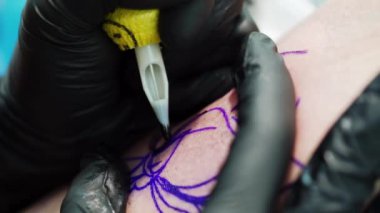 Ciltteki mavi resim dövme makinesinin siyah mürekkebiyle kaplanmış. Dövme sanatçısının eldivenli elleri beyaz deriyi geriyor. Kapat..