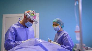 Renkli şapkalı cerrah tiroid ameliyatında çalışıyor. Doktor kullanılmış şırıngayı hemşireye verir ve yeni bir tane hazırlar..