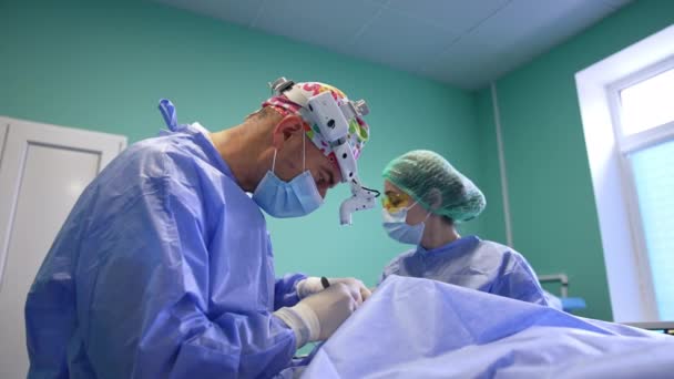 博士は慎重に切開するためにスカルペルを使用します 外科医を支援する黄色い保護ゴーグルの看護師 — ストック動画