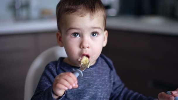 小さなコーカサス人の子供はフォークから自分自身を食べます 可愛い赤ちゃんがゆっくりと見上げています ブラーレッド バックドロップ — ストック動画