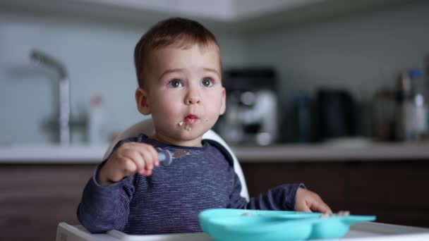 コーカサス トッドラーは 高い椅子に座って 徹底的に食べ物を噛んでいる 赤ん坊の少年はフォークで自分自身を食べることを学びます — ストック動画