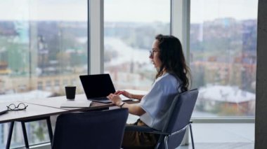 Dizüstü bilgisayarda çalışan genç bir bayan masasında oturuyor. Bağımsız yazar, gazeteci, metin yazarı. Arkaplanda bulanık şehir manzaralı panoramik pencereler.