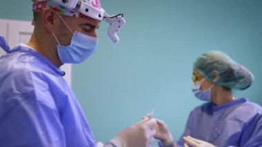 Kadın asistan bir doktora açık şırınga veriyor. Farlı bir cerrah hastanın yüzüne sünger sürer..