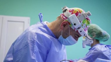 Komik şapkalı ve farlı bir erkek cerrah dikkatlice elektrikli cihaz kullanıyor. Kadın asistan doktor için makas ve ipliği uzattı.