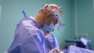 Renkli şapkalı, maskeli ve farlı kendine güvenen bir erkek cerrah meslektaşıyla konuşuyor. Doktor göz kapağı ameliyatı için aletler uyguluyor. Yan görünüm.