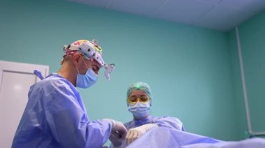 Erkek cerrah metal bir aletle ameliyat prosedürünü açıklıyor. Kadın doktor araç gereçleri arka planda ayarlar. Düşük açı görünümü.