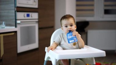 Beyaz, şirin bir erkek bebek yüksek bir sandalyede oturuyor. Toddler ağzında meyve püresi paketi tutuyor ve kameraya sevimli bir şekilde gülümsüyor. Bulanık arkaplan.