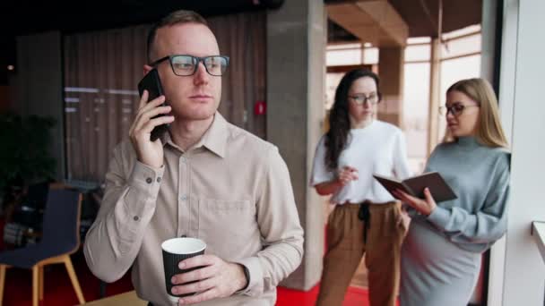 男人戴着眼镜 手里拿着咖啡杯 试着打电话 女人站在后台聊天 办公室午休 — 图库视频影像