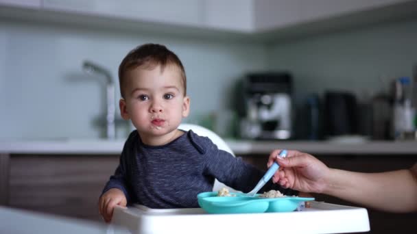 ママはスプーンからお粥で赤ちゃんに餌を与えています 子供の口の周りの食糧の部分 ブラーレッド バックドロップ — ストック動画