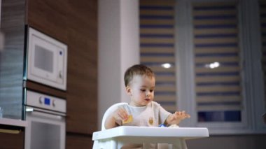 Yüksek sandalyedeki sevimli çocuk kurabiye ve çörek seçiyor. Bebek yemek için yemek seçiyor ve elinde bir çörek tutuyor..
