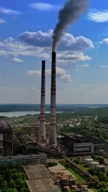 Doğa arasındaki sanayi bölgesi. Mavi gökyüzüne karşı boruları olan bir fabrika. Kara duman endüstriyel borudan çıkıyor. Doğanın arka planında üretimin panoramik görüntüsü. Dikey video