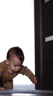 Kahverengi şortlu küçük bebek kapıya doğru sürünüyor. Güzel çocuk kapıları itiyor ve yere düşüyor. Dikey video