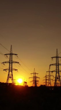 Gün batımında iletim kuleleri. Akşamları dumanı tüten endüstriyel pipoya yakın elektrik direkleri. Güneş batarken güzel turuncu gökyüzüne karşı yüksek voltajlı elektrik hatları. Enerji verimliliği kavramı. Dikey video