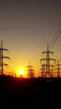 Elektrikli sütunlar turuncu günbatımına karşı. Akşamları yüksek voltajlı elektrik hatlarının koyu arkaplanı. Batan güneşte iletim hatları. Dikey video