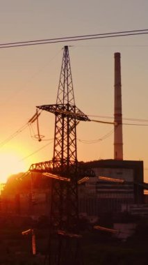Gün batımında elektrik direkleri. Endüstriyel boru arka planında yüksek voltajlı elektrik kulesi. Akşamları kirli dumanla fabrikanın yakınındaki iletişim hatları. Dikey video