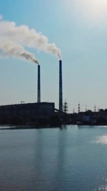 Gün içinde mavi gökyüzünün altında nehrin yanındaki büyük fabrikaya yakın hava manzarası. Göl kıyısında inşa edilmiş iki büyük borulu hidroelektrik santrali. Dikey video