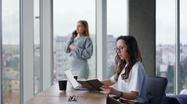 Dizüstü bilgisayarında uzun kıvırcık saçlı bir kadın elinde kağıt bir defter tutuyor. Sarışın hamile kadın pencerede dikilip telefonda konuşuyor..