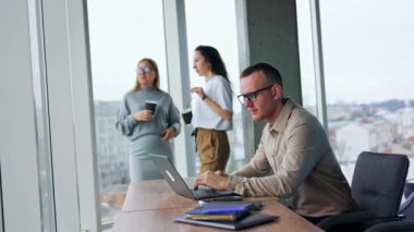 Erkek çalışan dizüstü bilgisayara odaklanmış. İşteyken sorun çözüyordum. Kadın iş arkadaşı sohbet ediyor ve arka planda kahve içiyor..