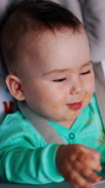 Tatlı küçük çocuk beslendikten sonra yemekle oynuyor. Sandalyede oturan sevimli bebek kaşığı ağzına alıyor. Kapatın. Dikey video