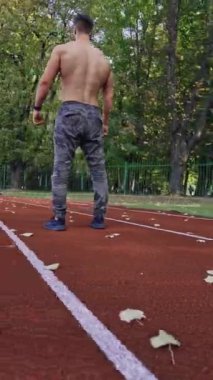 Spor koşucusu açık hava atletizm pistinde. Yeşil ağaçların arka planında stadyum yolunda koşan tişörtsüz bir sporcu. Hareket eden kamera var. Dikey video