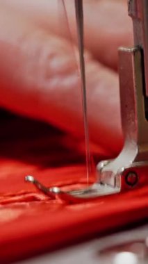 Kırmızı kumaşla çalışan dikiş makinesi. Kadının elleri, parlak kırmızı malzemeli metal dikiş makinesinde çalışıyor. Dikey videoyu kapat