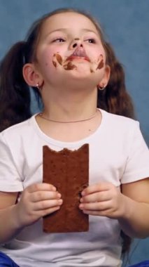 Çikolata yiyen edepsiz kız. Komik çocuk elinde çikolatayla stüdyoda izole edilmiş tatlı bir parçayı ısırıyor. Neşeli bir çocukluk. Dikey video