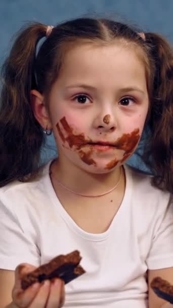 チョコレートを食べている女の子の汚い顔 子供の手にはチョコレートが2枚入っています スタジオで孤立した顔をした小さな子供の肖像 バーティカルビデオ — ストック動画