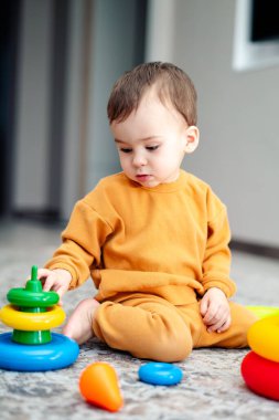 Bir bebek bir yığın blok ve bir oyuncakla oynuyor. Bebek sarı bir gömlek giyiyor ve yerde oturuyor.