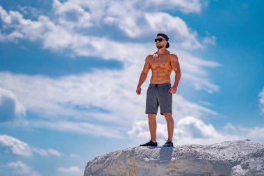 Kaslı vücutlu bir adam kollarını kavuşturmuş bir kayanın üzerinde duruyor. Şapka ve güneş gözlüğü takıyor..