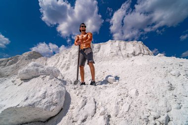 Bir adam elinde bir çubukla bir dağın tepesinde duruyor. Gökyüzü mavi ve arka planda bulutlar var..